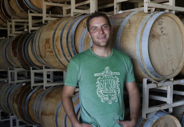 Anthony in a green T-shirt stands smiling in front of a stack of wooden barrels in the Sixmilebridge cellar or winery.