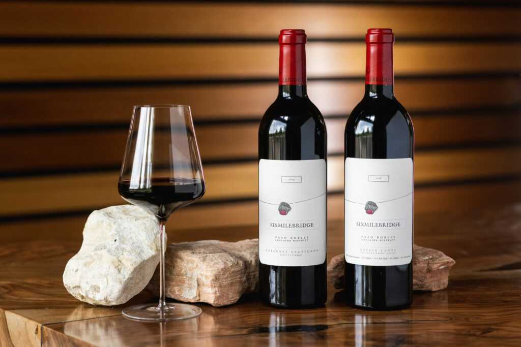 Two bottles of wine with labels and red caps are placed beside a wine glass filled with red wine. The setup includes decorative stones, and the background features horizontal wooden panels.