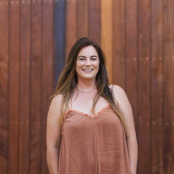 A woman with long brown hair stands smiling in front of a wooden wall. She is wearing a sleeveless brown top. The image has a warm and natural atmosphere.