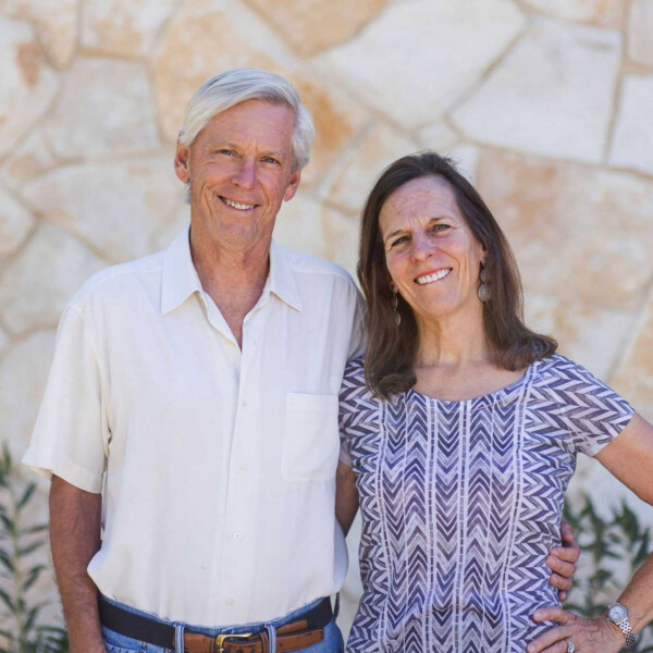 An older couple stands in front of a stone wall. The man, with gray hair, wears a white shirt and blue jeans. The woman, with long brown hair, wears a patterned dress. Both are smiling and appear relaxed.