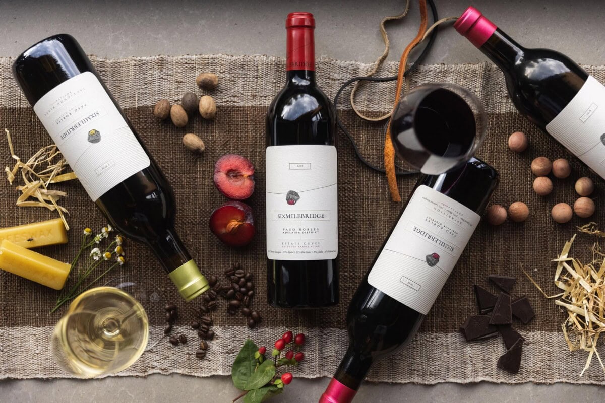 Four wine bottles are arranged on a textured brown cloth, surrounded by assorted items like a cut apple, nuts, chocolate, coffee beans, and herbs, creating a rustic and elegant display.
