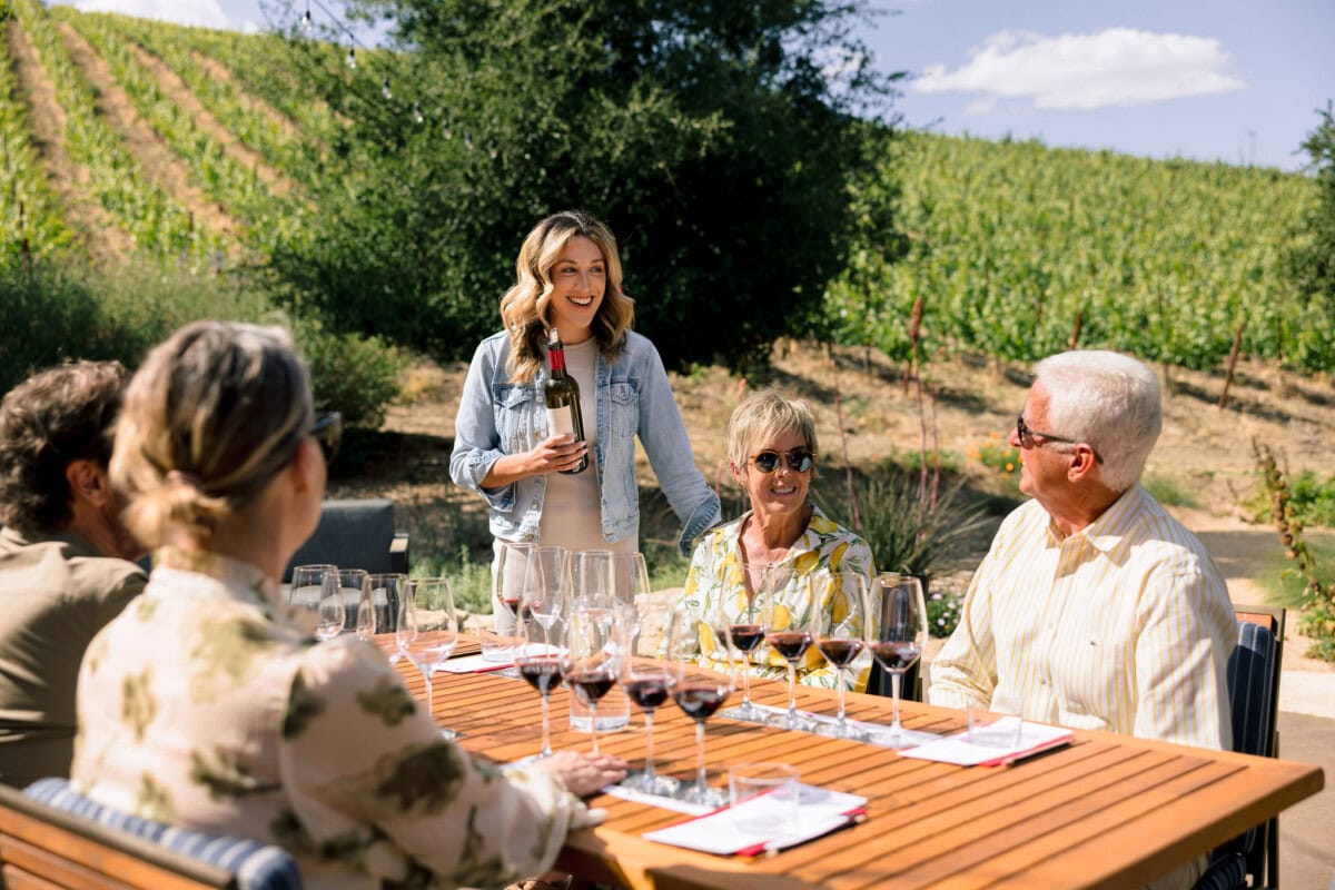 Woman serving wine to two men and two women. Table is set for a tasting. Vineyards in the background.