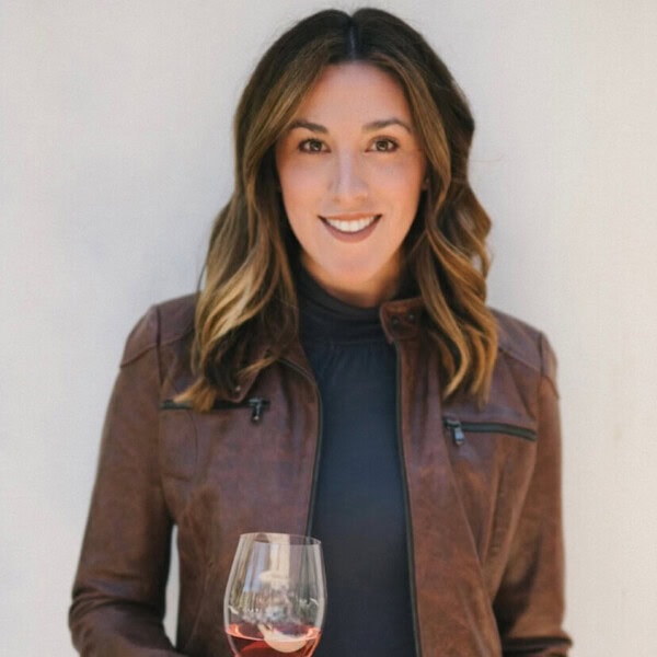 A woman with long hair wearing a brown leather jacket and jeans holds a glass of pink wine in one hand and a wine bottle in the other. She stands against a light-colored background, smiling at the camera.