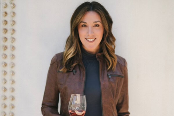 A woman with long hair wearing a brown leather jacket and jeans holds a glass of pink wine in one hand and a wine bottle in the other. She stands against a light-colored background, smiling at the camera.