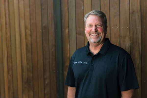 Man in black shirt smiling at camera and standing in front of wood wall.