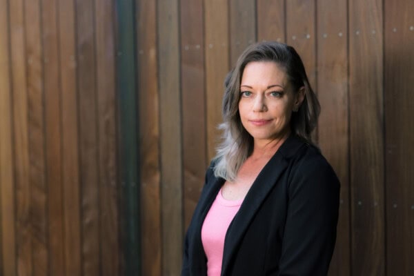 Woman in black jacket and pink shirt standing in front of wood wall.