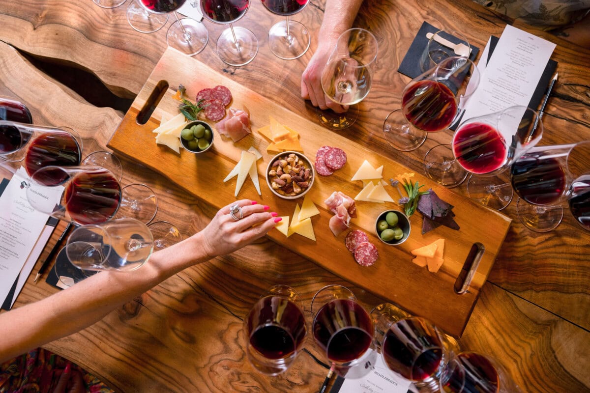 Table set with a dozen glasses of wine and charcuterie platter. Woman's hand reaches for cheese, man reaches for glass of wine.
